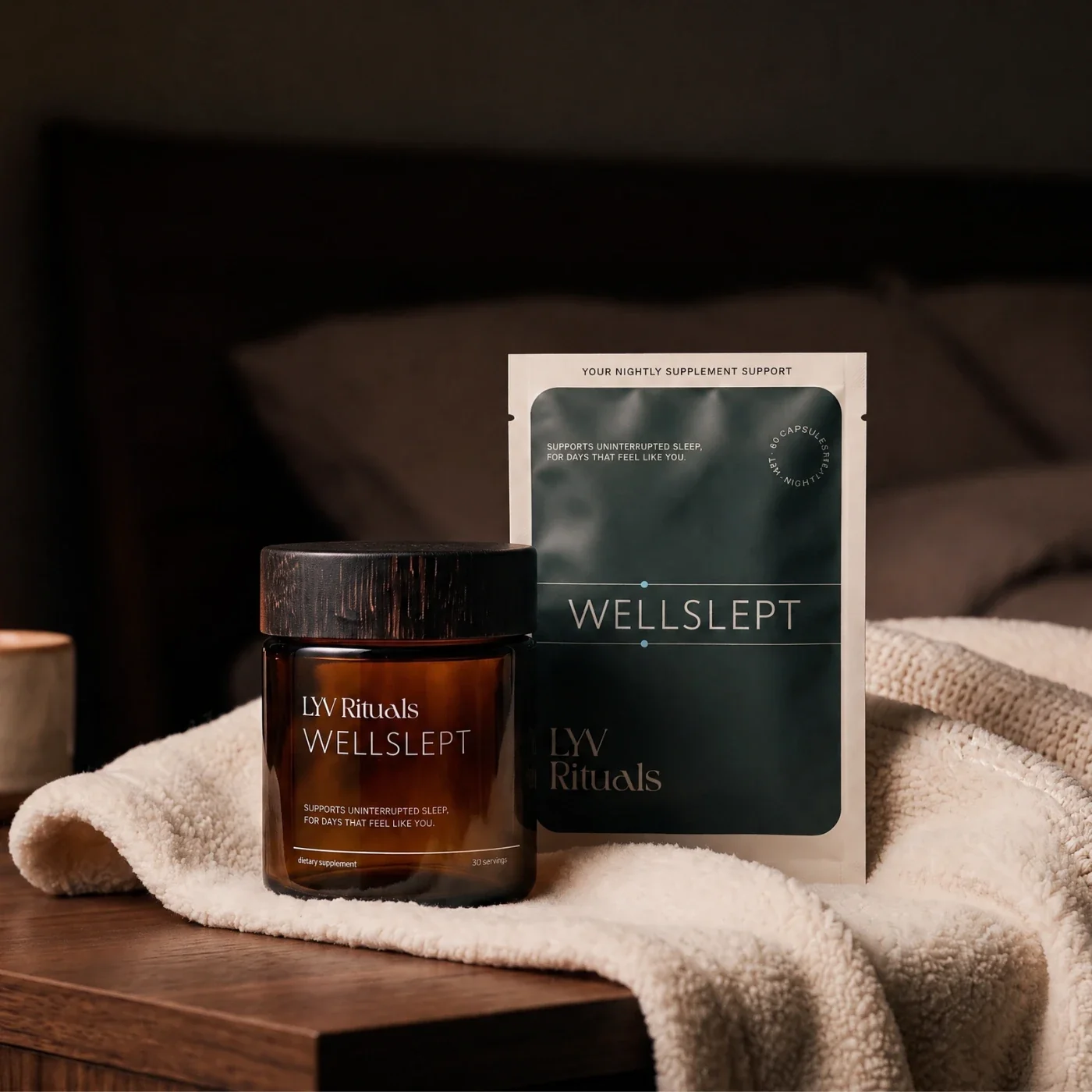 WELLSLEPT amber glass jar with a wooden lid next to the WELLSLEPT refill pouch, on a cream blanket on a bedside table